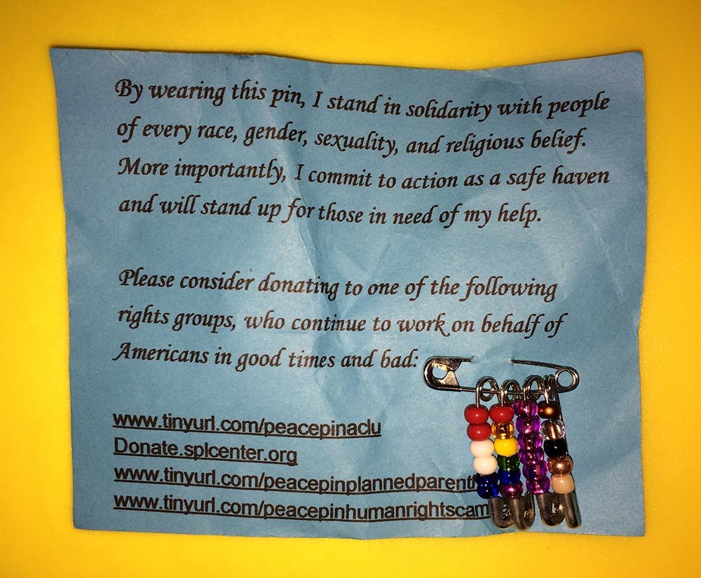Safety pins with colorful beads on them, attached to a card encouraging donations