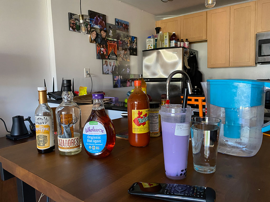 A view of my kitchen from the living room. A wall of large photos of family and friends. A table with a water jug, a purple boba tea, and other condiments.