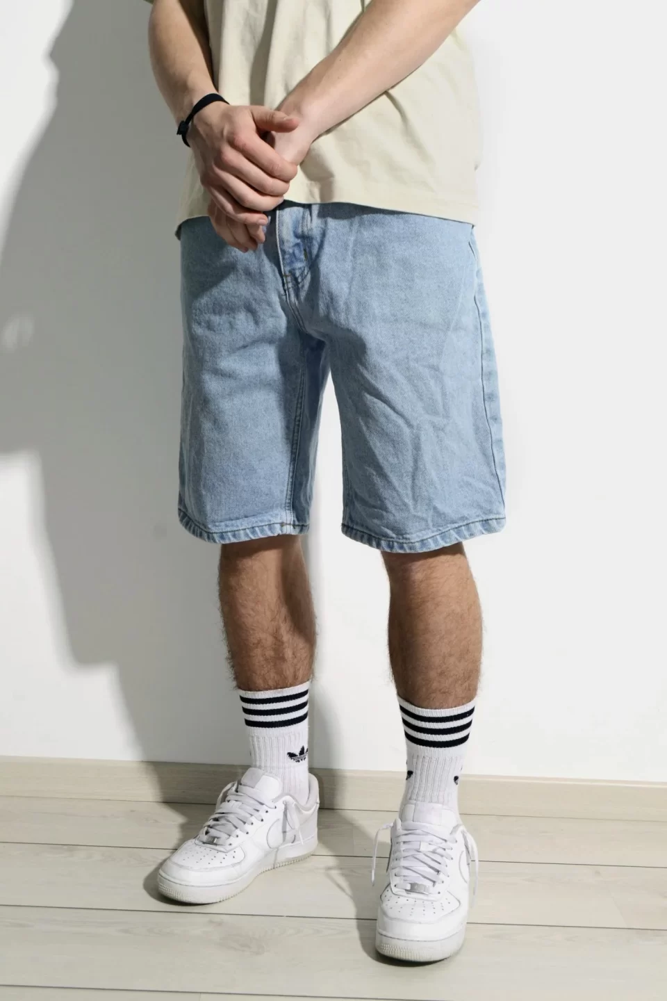 Denim shorts long enough to land beneath the knee of the model who stands awkwardly in a pale tan shirt and knee high striped white socks with white sneakers.