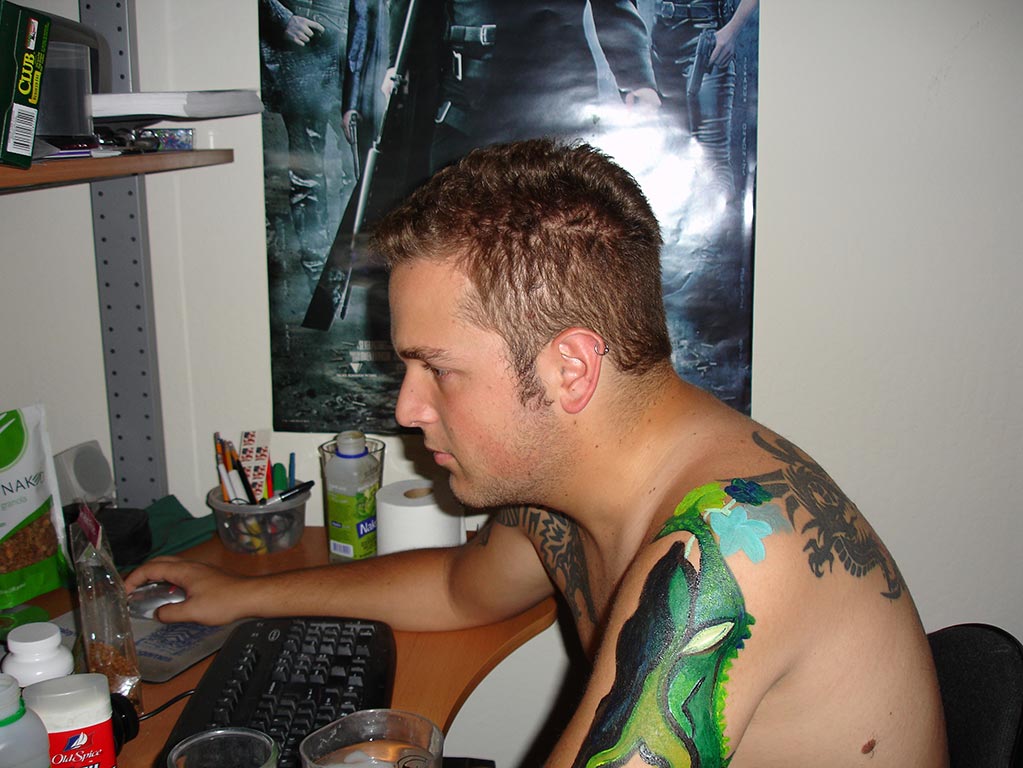 Candid 2007 photo of Corry sitting at his desk at home in the room he was renting. The desk is covered in food, toiletries, and other supplies indicating how small a living space this was. The Matrix movie poster is on the wall behind him. His arm is painted green with an acrylic paint design by Kati who took the photo.