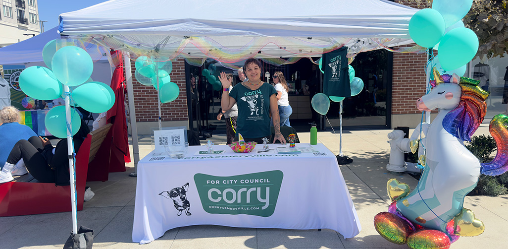 Corry waving from her table at Emeryville Pride 2026 at Bay Street