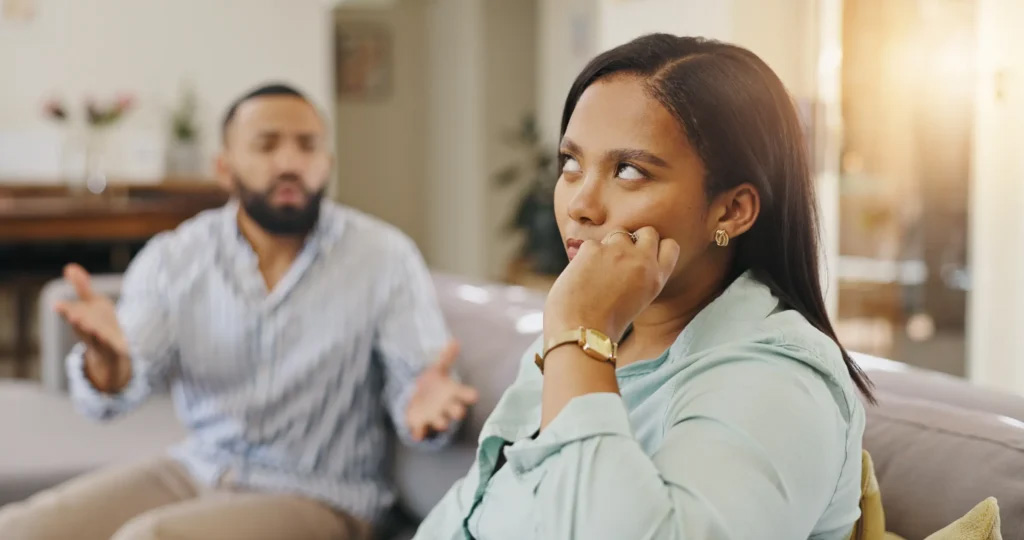 A woman rolls her eyes as her partner seems exasperated on the couch next to her