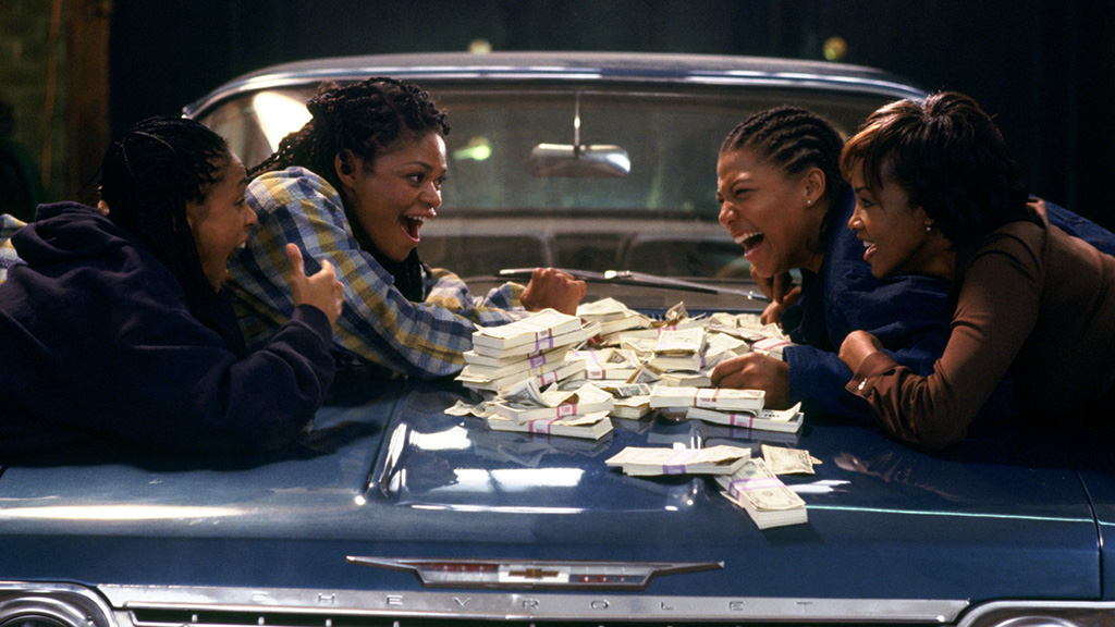 Four black women excitedly lean over a big pile of cash on the hood of a blue Chevy.
